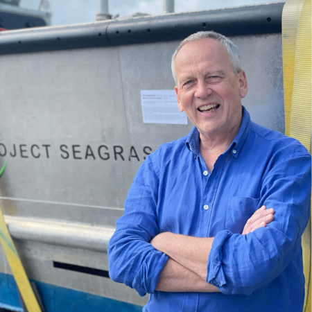 Rob Raimes is standing next to the Project Seagrass boat. He is wearing a blue shirt and has his arms folded in front of him.
